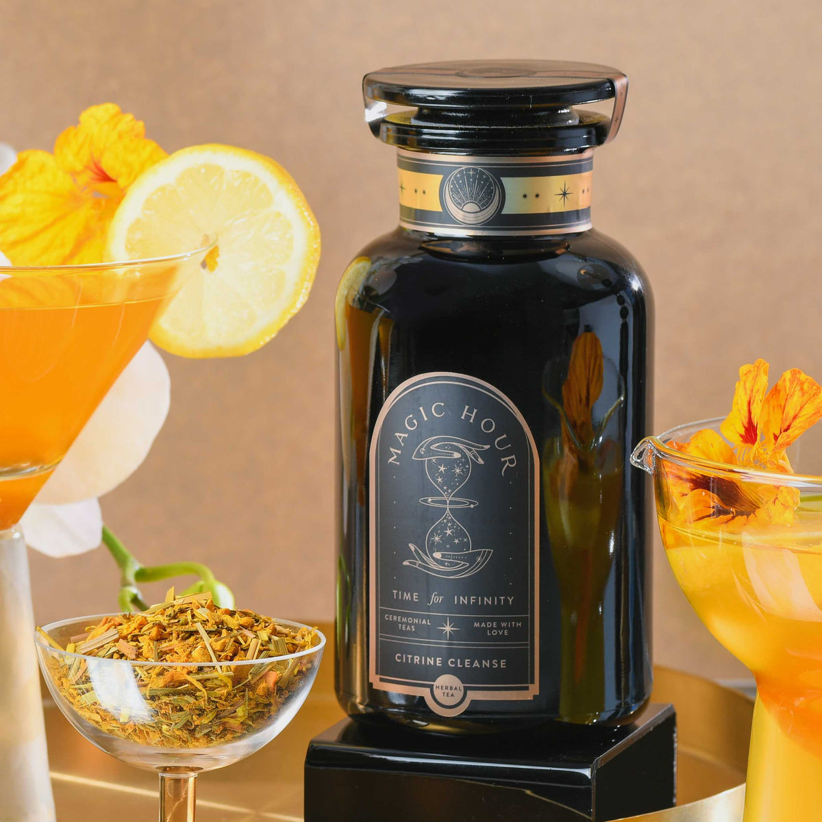 A black tea jar labeled “Magic Hour” sits among orange cocktails, a lemon slice, yellow flower petals, and loose tea—perhaps a detoxifying elixir of Citrine Cleanse™ Herbal Tea—on a gold tray, against a warm beige background.