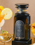 A black tea jar labeled “Magic Hour” sits among orange cocktails, a lemon slice, yellow flower petals, and loose tea—perhaps a detoxifying elixir of Citrine Cleanse™ Herbal Tea—on a gold tray, against a warm beige background.