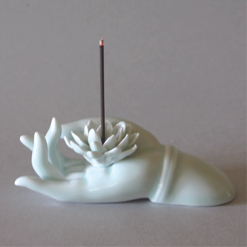 The Hand Lotus Incense Stand by Eastern Elm is a minimalist piece of home decor designed to resemble a graceful hand, crafted from smooth white material. The open hand cradles a lotus flower, where an incense stick can be inserted and lit, emitting a delicate wisp of smoke—perfect for your meditation practice.