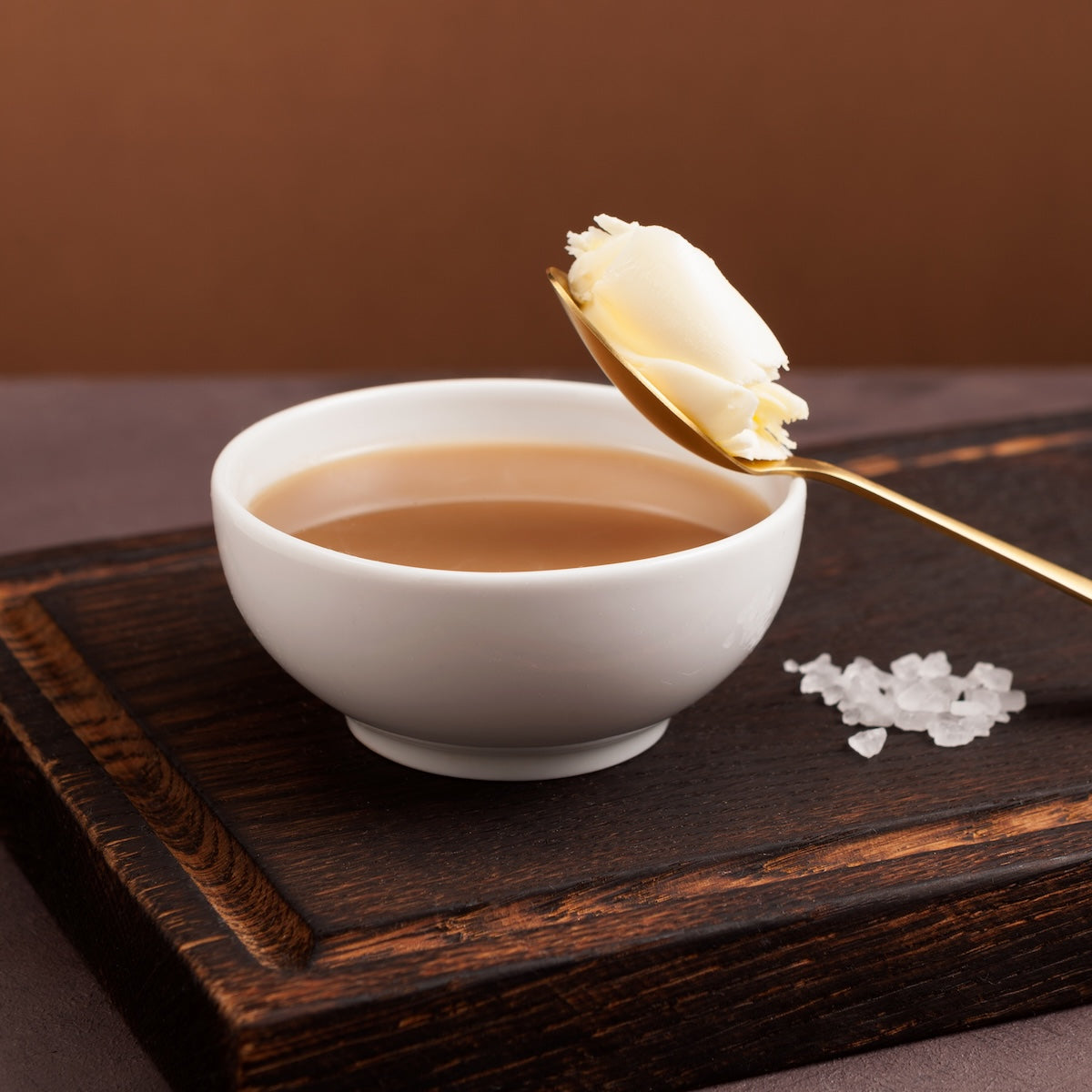 A white bowl of Butter Puerh sits on a dark wooden board with a spoon holding a dollop of butter above. Coarse salt crystals are scattered nearby on the warm brown surface.