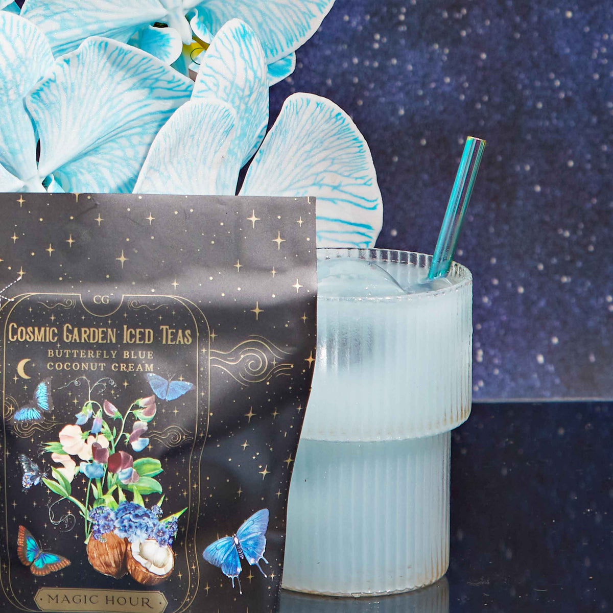 A glass of light blue iced tea with the Aurora Bombilla stainless steel tea straw sits beside a decorative tea package and blue and white flowers, set against a starry night background.