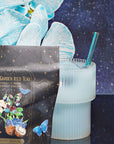 A glass of light blue iced tea with the Aurora Bombilla stainless steel tea straw sits beside a decorative tea package and blue and white flowers, set against a starry night background.