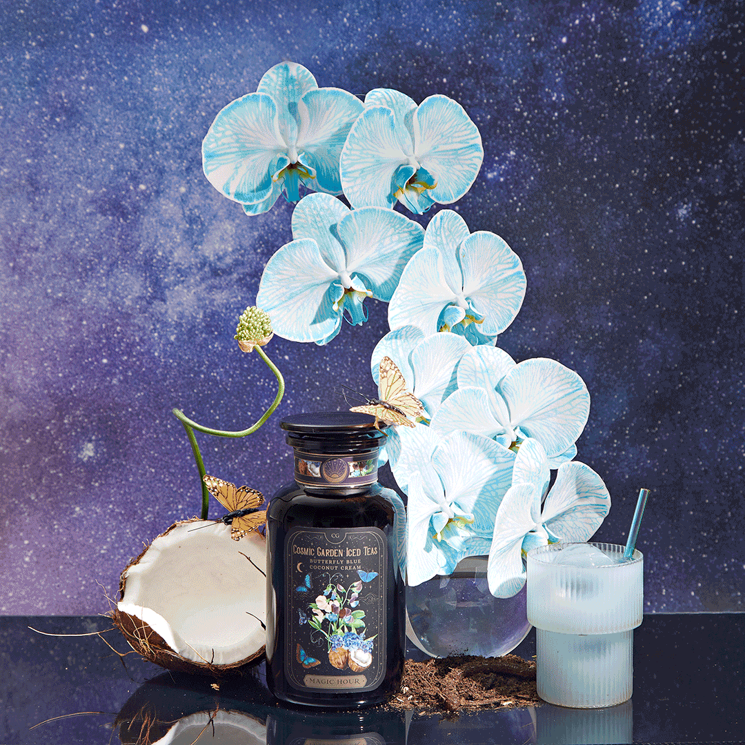 A dark jar labeled "Butterfly Blue Coconut Cream Iced White Tea" sits beside a halved coconut, a glass of milk with a straw, and blue pea flower orchids against a starry galaxy background. Small butterflies rest on the flowers.