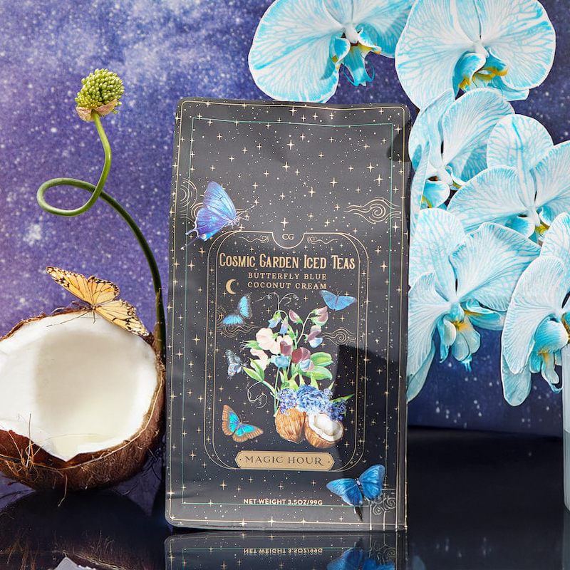 A package of Butterfly Blue Coconut Cream Iced White Tea Refill Pouch stands out against a starry sky. Its celestial design pairs with blue orchids and half a coconut, perfect for refreshment this summer.