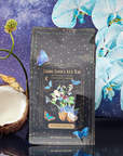 A package of Butterfly Blue Coconut Cream Iced White Tea Refill Pouch stands out against a starry sky. Its celestial design pairs with blue orchids and half a coconut, perfect for refreshment this summer.