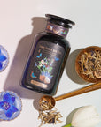 A glass jar labeled "Magic Hour Butterfly Blue Coconut Cream Iced White Tea" is surrounded by two glass cups with blue flowers, a gold spoon with loose leaf tea, and a white tulip. The golden spoon is engraved with "Magic Hour," evoking the perfect moment for summer hydration. The scene is lit with soft, natural light.