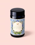 A dark jar labeled "Flower Powder: Butterfly Bergamot Matcha," adorned with blue floral designs, rests against a pastel pink background. This exquisite blend unites ceremonial matcha with butterfly pea flower and vibrant notes of bergamot to create a truly enchanting tea experience.