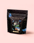A pouch labeled "Butterfly Blue Coconut Cream Iced White Tea" shows blue butterflies, gold stars, blue flowers, a coconut, and green leaves on the front against a pale pink background, hinting at pina colada flavor.
