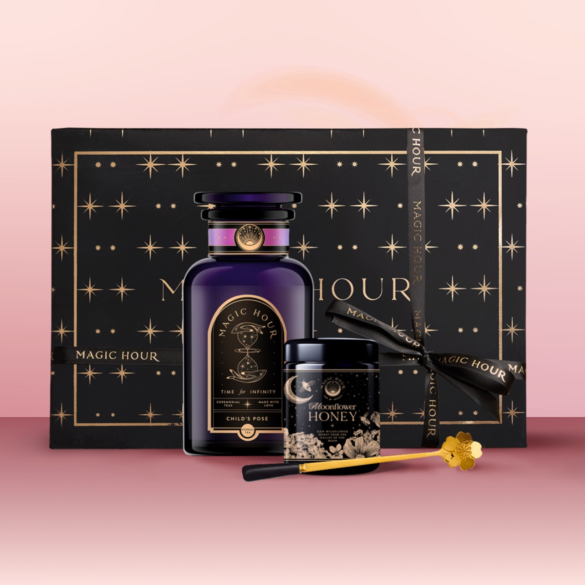 The Tea &amp; Honey Gift Set features a black and gold star-patterned box, dark purple &quot;Magic Hour&quot; bottle, wildflower honey, and a golden honey dipper—all arranged on a pink background for your perfect morning ritual.