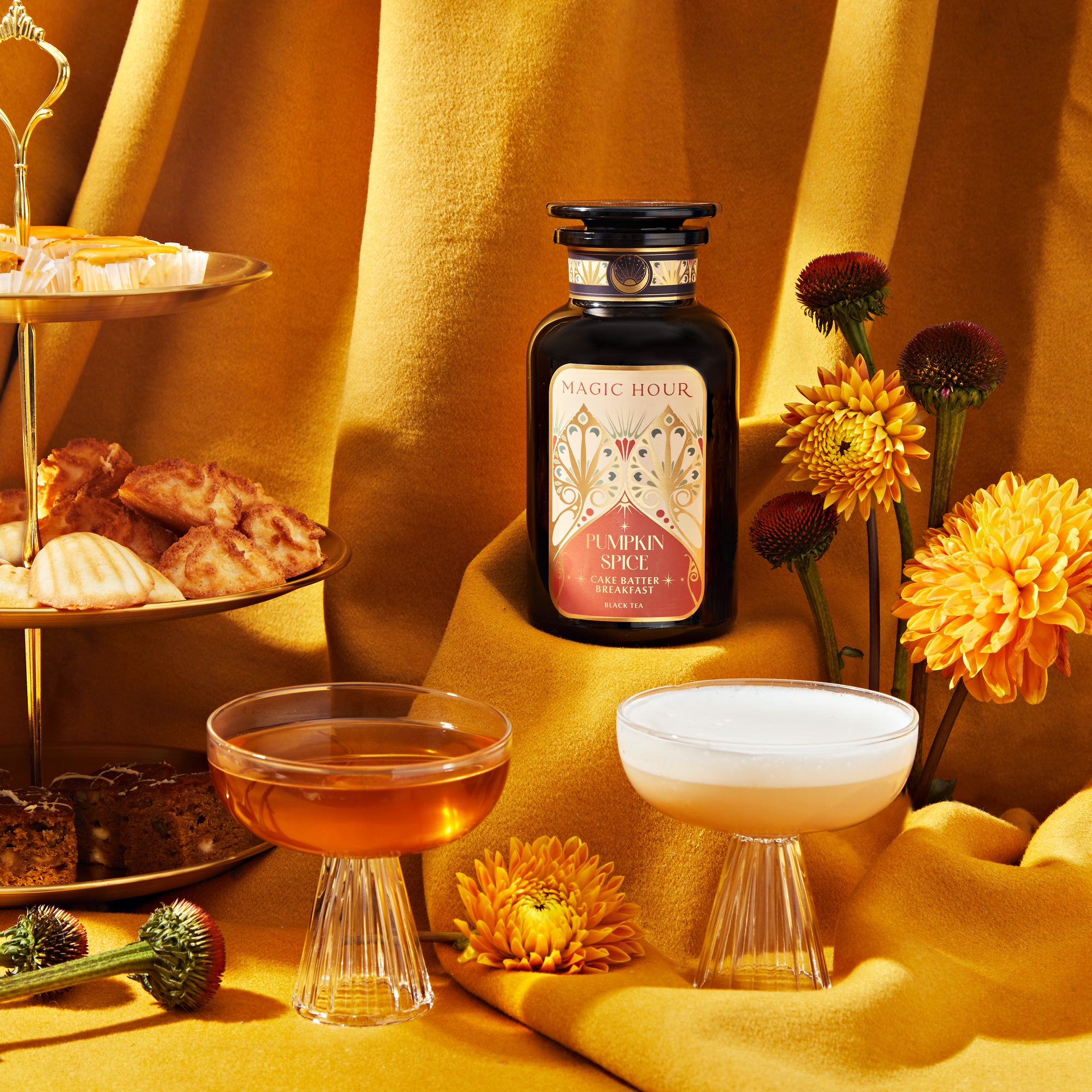 A bottle from the Pumpkin Spice Essentials Tea Trio Gift Set is showcased on a golden cloth, flanked by two glass cups filled with comforting brew. An assortment of pastries and vibrant yellow flowers, along with apothecary jars, adds warmth and elegance to this enchanting scene.