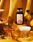 A bottle from the Pumpkin Spice Essentials Tea Trio Gift Set is showcased on a golden cloth, flanked by two glass cups filled with comforting brew. An assortment of pastries and vibrant yellow flowers, along with apothecary jars, adds warmth and elegance to this enchanting scene.