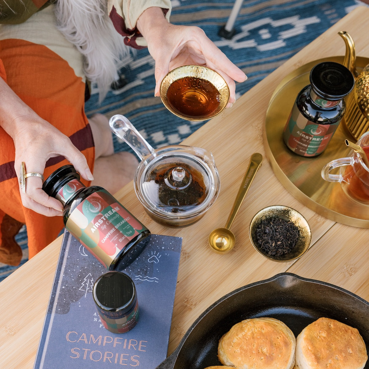 Campfire Breakfast Tea in various sizes on a wooden table with a campfire stories book, cast iron pan with biscuits, and a gold cup with loose leaf tea. 