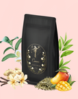A black bag labeled "Cancer: Queen of Cool Mango-Ginger-Cream White Tea" sits on a light pink background, surrounded by illustrations of organic ginger, mango, vanilla, tea leaves, and white flowers.