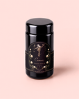 A black jar labeled “Cancer: Queen of Cool Mango-Ginger-Cream White Tea,” adorned with gold floral designs and an illustrated figure, set against a pale pink background, evokes elegance while hinting at the nourishing benefits of organic mango and ginger.