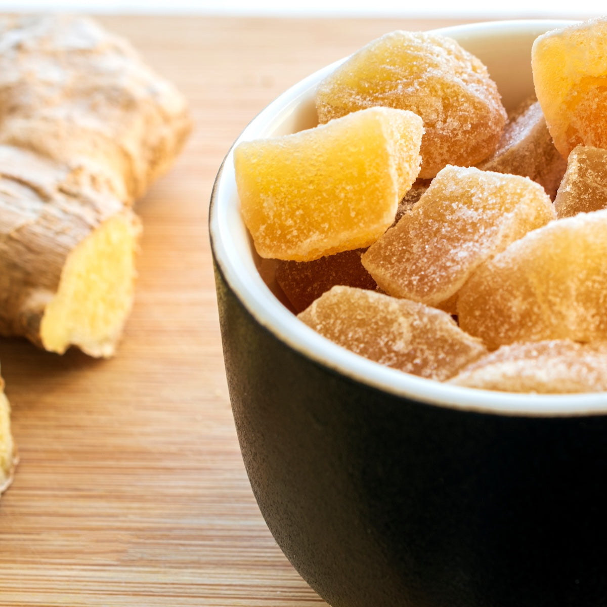 A bowl filled with sugar-coated pieces of Ginger Lemon sits on a wooden surface, with a piece of fresh ginger root in the background.