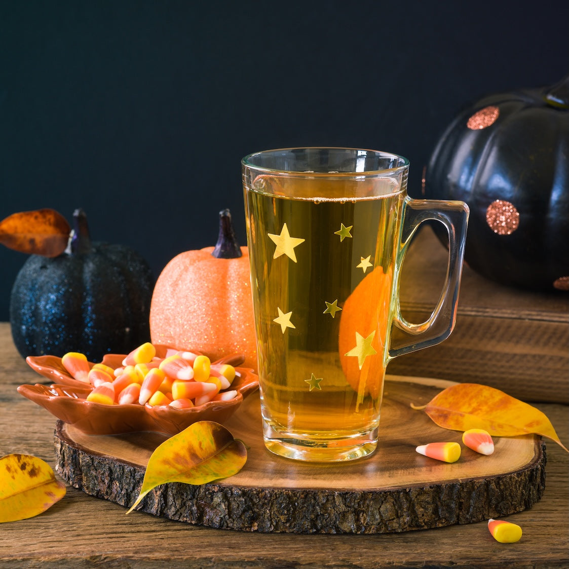 A glass mug with star designs holds yellow Candy Corn tea, set on a wooden slab beside a dish of Candy Corn, autumn leaves, and glittery black and orange pumpkins for a festive Halloween display.