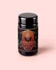 A black tea canister with a gold and red label reads “Candy Corn: Herbal Fall Tea with Vanilla & Caramel.” The design showcases celestial and mystical imagery on a light pink background, making this caffeine-free herbal tea blend the perfect Halloween treat for cozy autumn evenings.