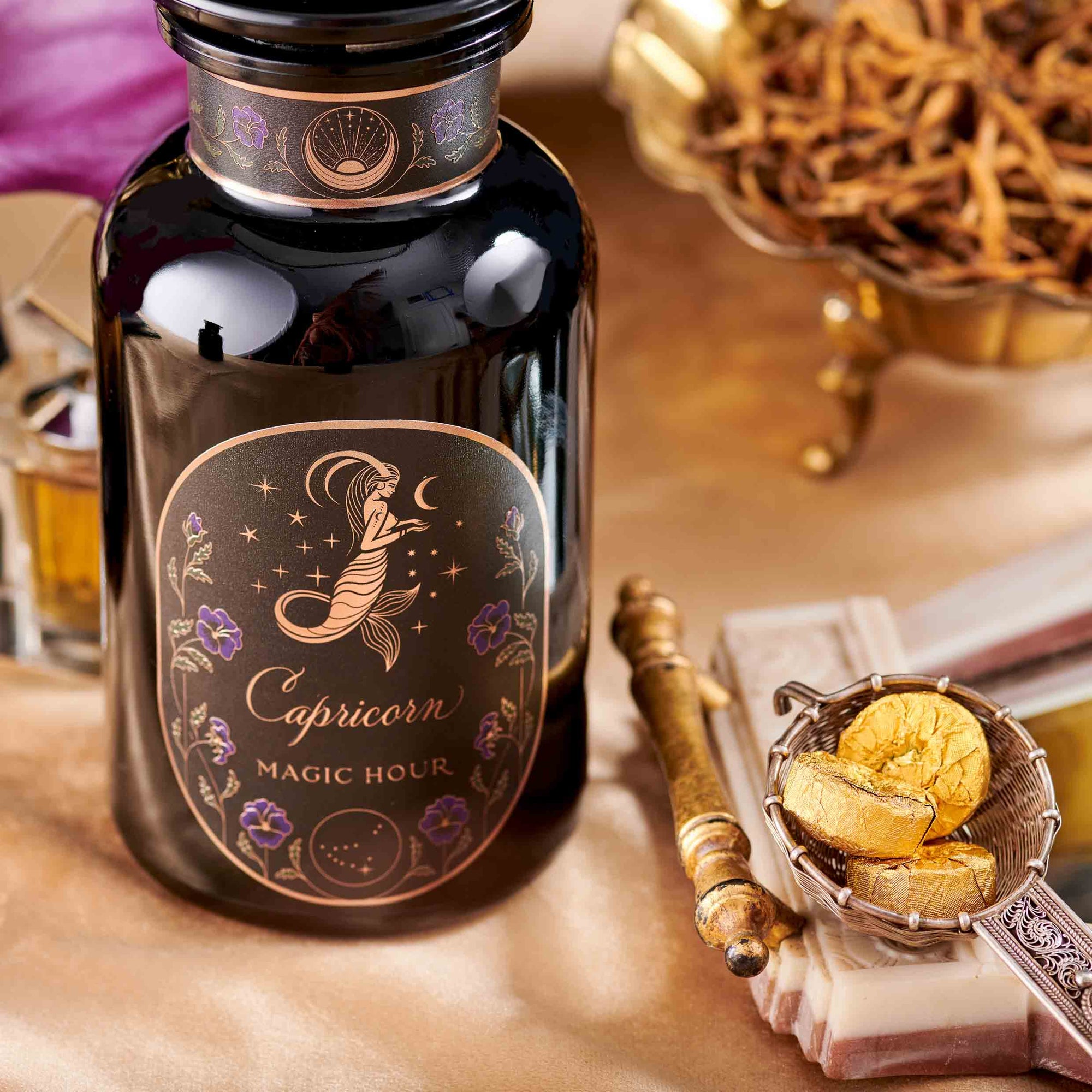 A black glass bottle labeled "Capricorn: Maple Oolong Tea" displays a mystical mermaid and celestial design, surrounded by gold objects and dried herbs, hinting at the tea's metabolism-boosting magic on an elegant surface.