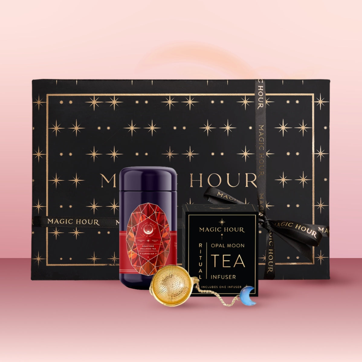 The Carnelian July Birthstone Mini Gift Set features a star-patterned black box, a red and gold Carnelian Wellness Tea tin, a gold tea infuser, and an Opal Moon Tea Infuser, displayed on pink.