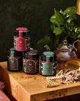 Four decorative jars labeled as Ceremonial Bath Soaks are stacked on a wooden table, accompanied by a plant, glass teapot, and golden scoop spilling colorful soaks. A tassel and beaded necklace enhance the elegance of this cosmic connection.