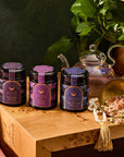 Three tea containers labeled "Sunday Saturn," "Friday Venus," and "Wednesday Mercury" sit on a wooden table, reflecting the celestial healing of Ceremonial Bath Soaks. Nearby, a glass teapot and loose tea leaves are ready, with a leafy plant adding greenery in the background.