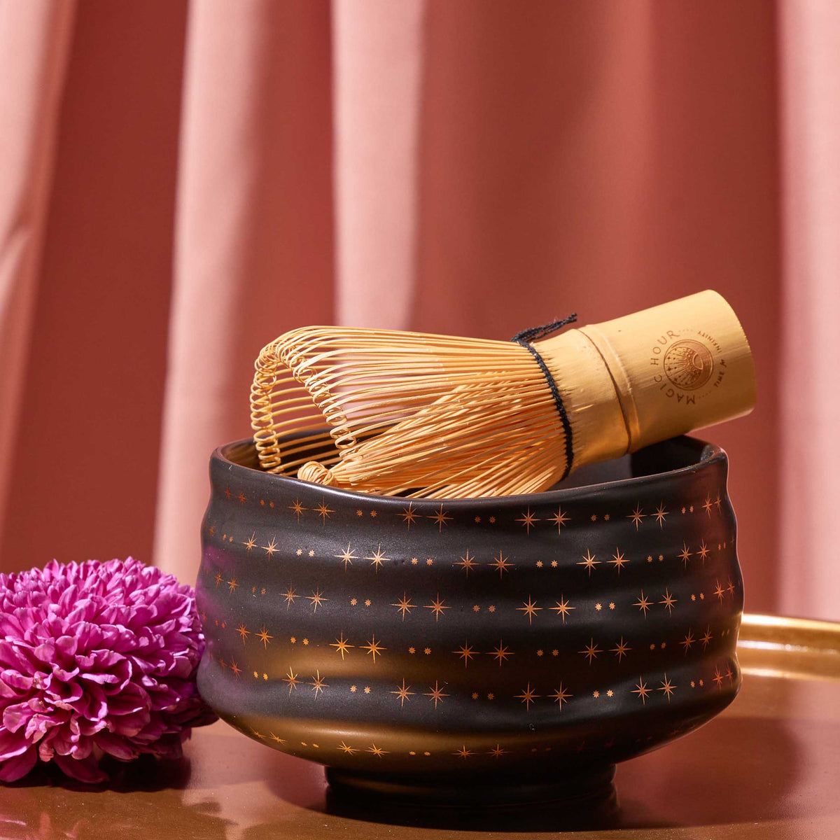 A bamboo matcha whisk rests on the Ceremonial Matcha Bowl, a handmade ceramic piece adorned with delicate 14k gold detailing and star patterns. A purple chrysanthemum flower sits nearby, while pink curtains form the background.