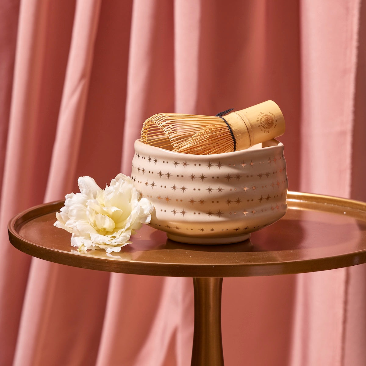 A cream-colored, star-patterned ceramic bowl from the Cozy & Connected Bundle with a bamboo whisk inside rests on a round, gold tray. A white flower lies beside the bowl. The background features pink, pleated fabric drapes.