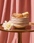 A cream-colored, star-patterned ceramic bowl from the Cozy & Connected Bundle with a bamboo whisk inside rests on a round, gold tray. A white flower lies beside the bowl. The background features pink, pleated fabric drapes.