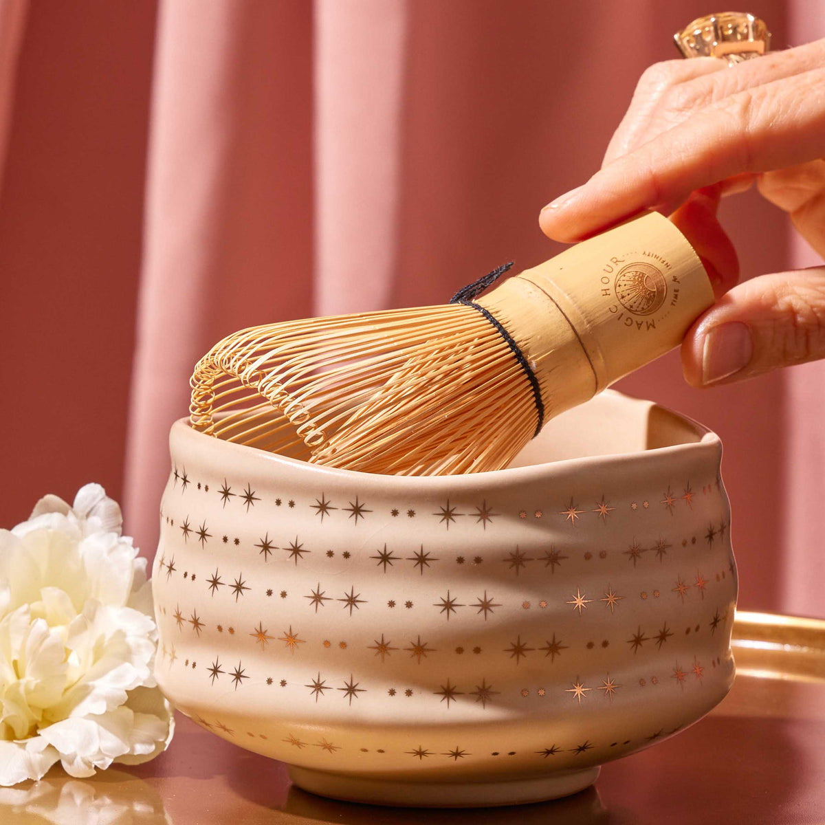 A hand holds a bamboo matcha whisk over the Ceremonial Matcha Bowl, which is handmade and features star patterns, beautifully crafted using traditional ceramic techniques. A white flower and a pink curtain appear in the background.