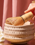 A hand holds a bamboo matcha whisk over the Ceremonial Matcha Bowl, which is handmade and features star patterns, beautifully crafted using traditional ceramic techniques. A white flower and a pink curtain appear in the background.