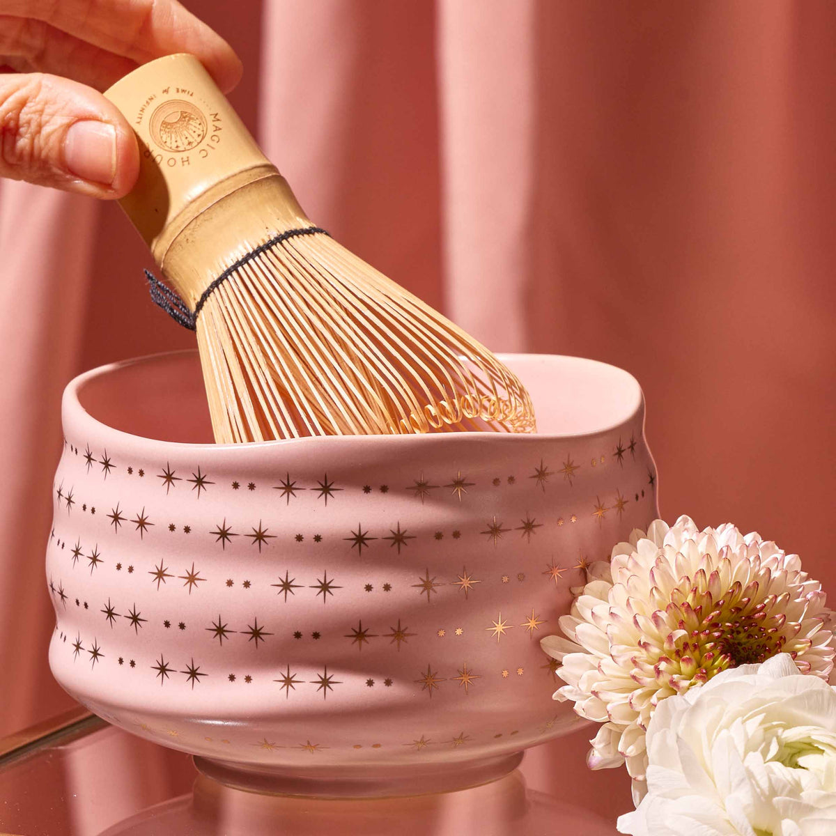 A hand holds a bamboo whisk over the Ceremonial Matcha Bowl, a handmade ceramic piece adorned with star patterns and delicate 14k gold detailing, displayed next to white flowers against a blush pink background.