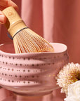 A hand holds a bamboo whisk over the Ceremonial Matcha Bowl, a handmade ceramic piece adorned with star patterns and delicate 14k gold detailing, displayed next to white flowers against a blush pink background.