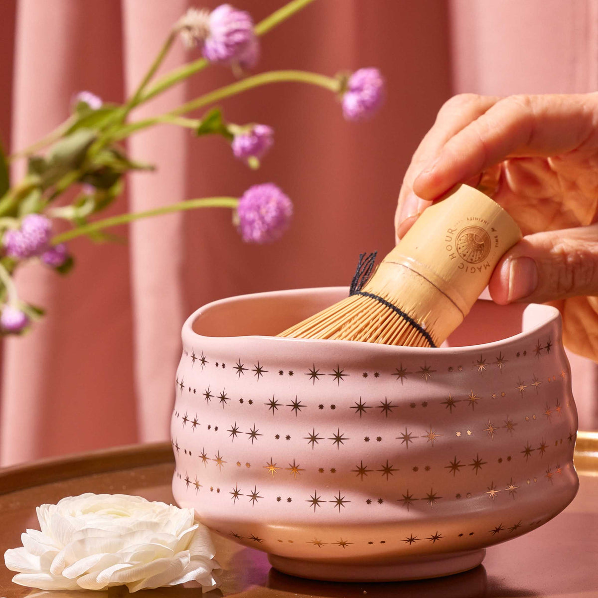 A hand holds a bamboo whisk inside the Ceremonial Matcha Bowl, a handmade ceramic piece featuring 14k gold detailing and delicate star patterns. Nearby, a white flower and purple flowers rest on a pink background.