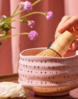 A hand holds a bamboo whisk inside the Ceremonial Matcha Bowl, a handmade ceramic piece featuring 14k gold detailing and delicate star patterns. Nearby, a white flower and purple flowers rest on a pink background.