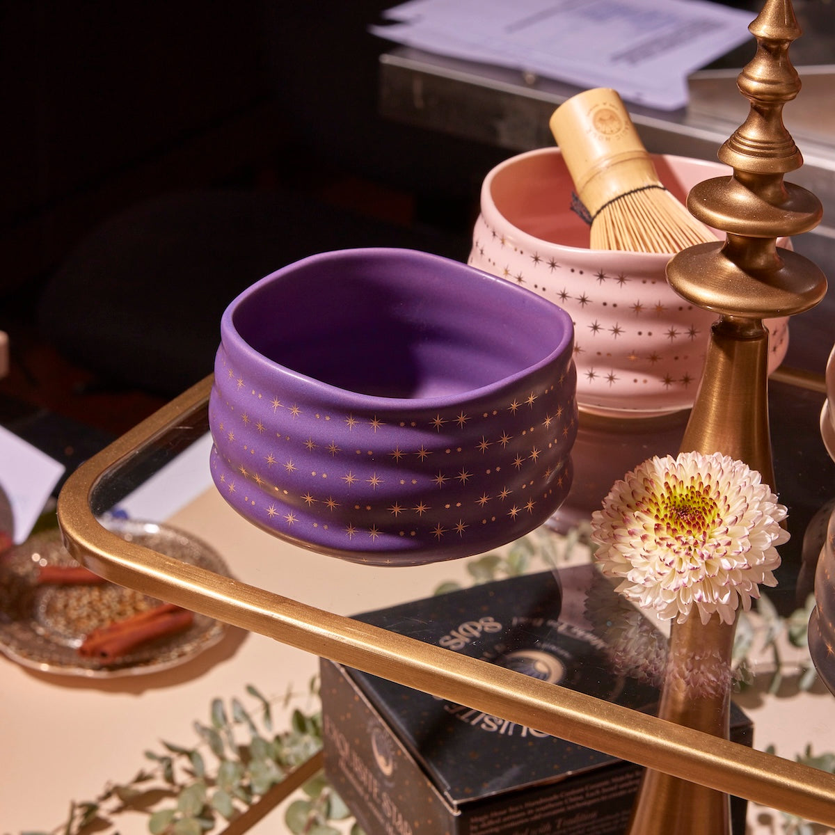 An Amethyst Gemstone Tea Gift Set bowl, adorned with gold stars on a purple ceramic background, rests on a glass table beside a pink bowl holding a bamboo whisk. In the background are a decorative stand, a white flower, and various papers and items.