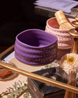 An Amethyst Gemstone Tea Gift Set bowl, adorned with gold stars on a purple ceramic background, rests on a glass table beside a pink bowl holding a bamboo whisk. In the background are a decorative stand, a white flower, and various papers and items.