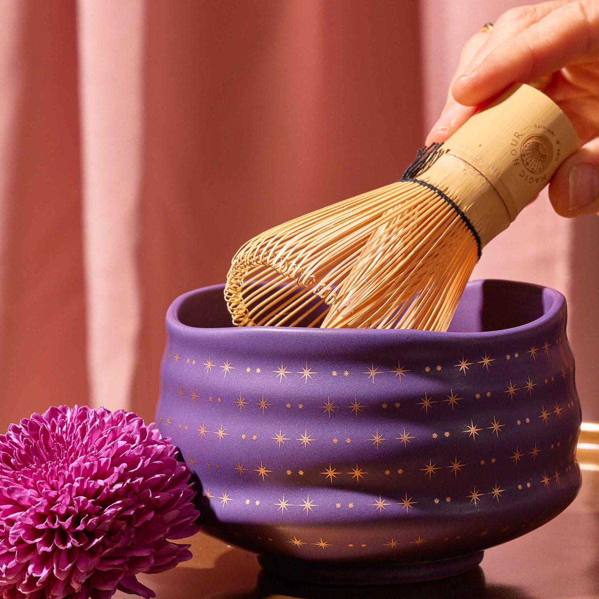 A hand holds a bamboo matcha whisk over the Ceremonial Matcha Bowl, a handmade ceramic piece adorned with shimmering gold star patterns reminiscent of 14k gold detailing. Beside the bowl rests a purple flower, set against the gentle backdrop of a soft pink curtain.