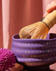 A hand holds a bamboo matcha whisk over the Ceremonial Matcha Bowl, a handmade ceramic piece adorned with shimmering gold star patterns reminiscent of 14k gold detailing. Beside the bowl rests a purple flower, set against the gentle backdrop of a soft pink curtain.