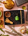 A Ceremonial Matcha Traveler Gift Box with organic ceremonial matcha, a bamboo whisk, and a copper mesh strainer, surrounded by a gold teapot, bowls of prepared tea, and a tea light in a crystal candle holder.