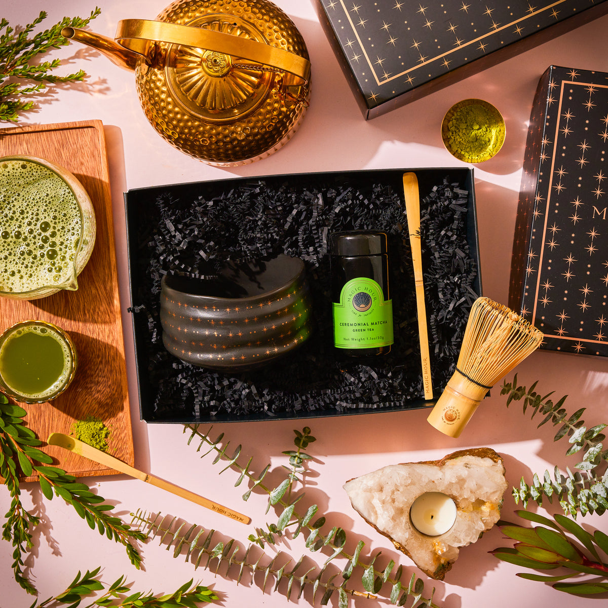 A Ceremonial Matcha Traveler Gift Box containing a black tea bowl, a tin of organic ceremonial matcha, a bamboo whisk, and a scoop, arranged with a gold teapot, glass pitcher, and crystal candle holder on a pink surface.