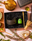 A Ceremonial Matcha Traveler Gift Box containing a black tea bowl, a tin of organic ceremonial matcha, a bamboo whisk, and a scoop, arranged with a gold teapot, glass pitcher, and crystal candle holder on a pink surface.