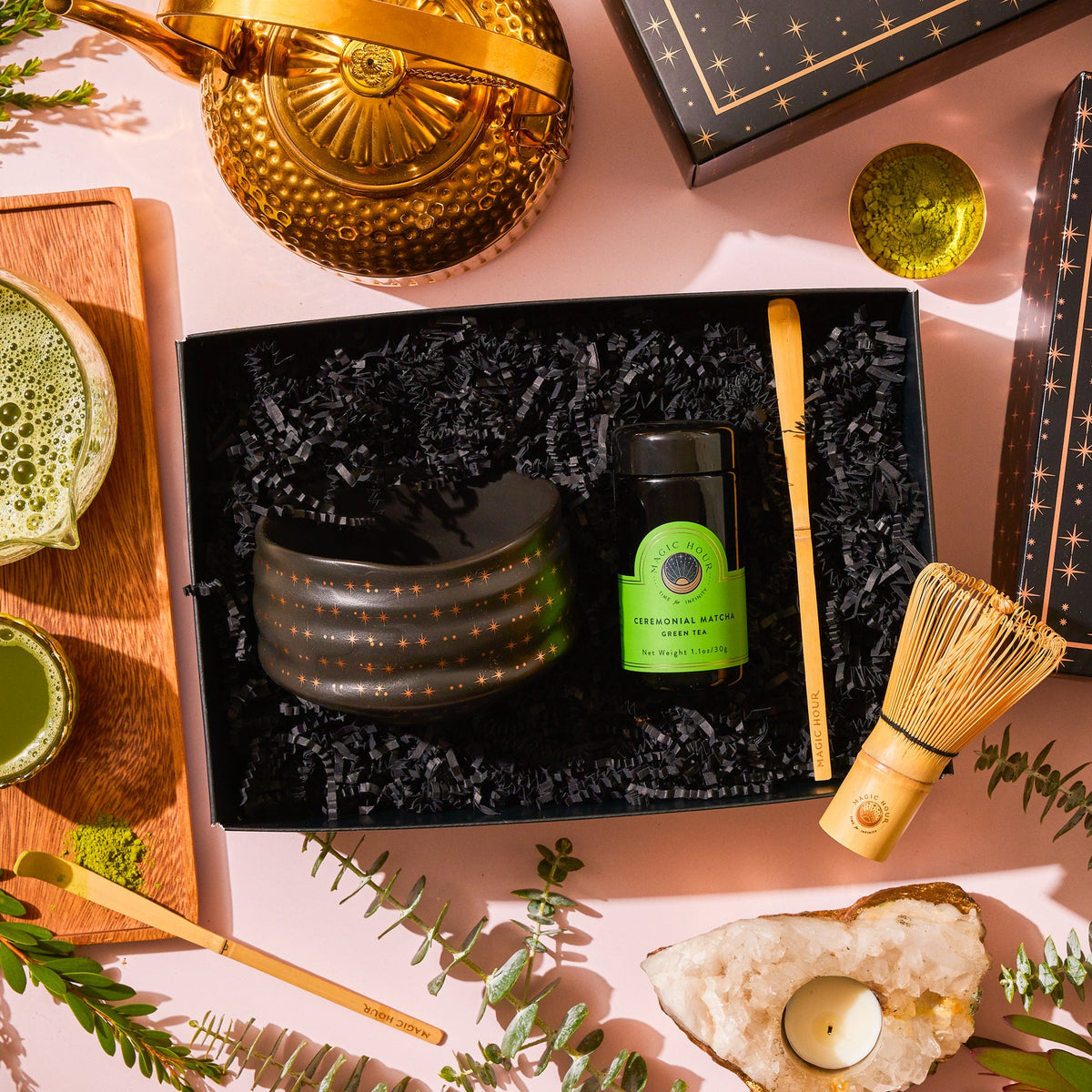 A Matcha Ceremony Gift Set in a box with a dark tea bowl, ceremonial matcha, and bamboo whisk, surrounded by a gold kettle, tea cups on a wooden board, and a tea light in a crystal holder.