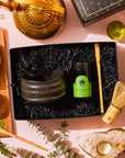 A Matcha Ceremony Gift Set in a box with a dark tea bowl, ceremonial matcha, and bamboo whisk, surrounded by a gold kettle, tea cups on a wooden board, and a tea light in a crystal holder.