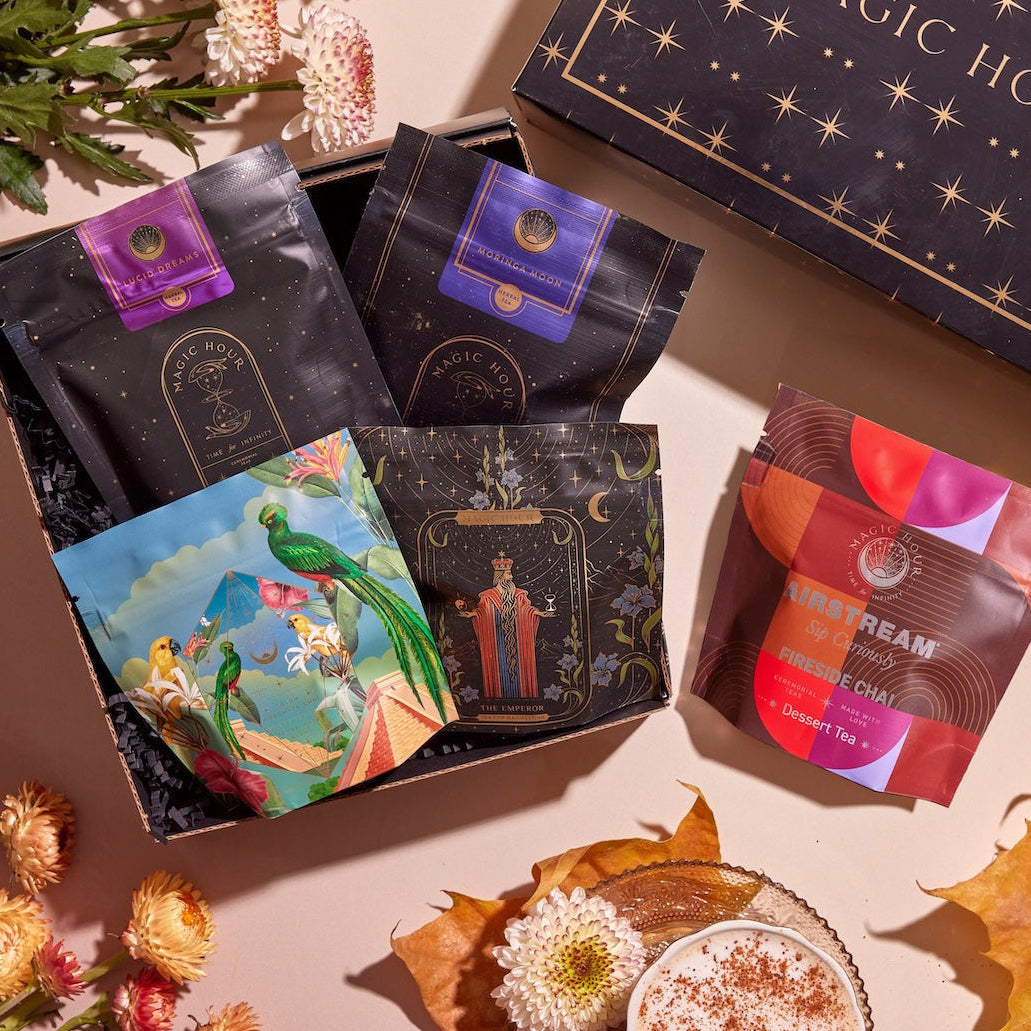 A beautifully styled arrangement featuring a decorative box labeled "Magic Hour" with four vibrant Chai Sampler packages inside. Surrounding the Chai Sampler are autumn leaves, flowers, and a cup of frothy chai tea with sprinkled spices, set on a pinkish background.