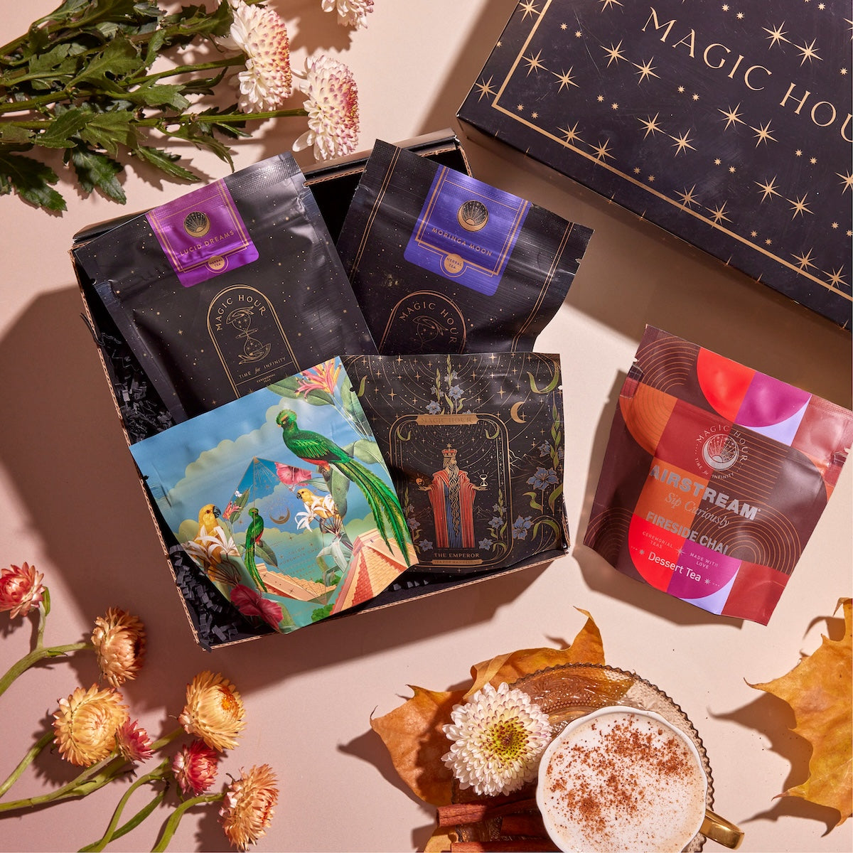 A beautifully styled arrangement featuring a decorative box labeled &quot;Magic Hour&quot; with four vibrant Chai Sampler packages inside. Surrounding the Chai Sampler are autumn leaves, flowers, and a cup of frothy chai tea with sprinkled spices, set on a pinkish background.