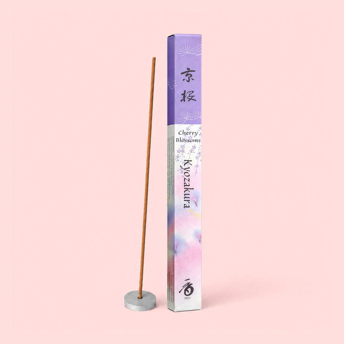 An incense stick infused with Kyoto Cherry Blossoms stands upright in a small holder beside a decorative box adorned with purple and white floral designs labeled “Natural Incense : Kyoto Cherry Blossoms.” The delicate fragrance of sakura cherry blossoms fills the air, evoking the gentle scents of springtime. The background is a pale pink shade.