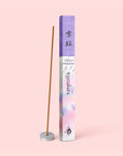 An incense stick infused with Kyoto Cherry Blossoms stands upright in a small holder beside a decorative box adorned with purple and white floral designs labeled “Natural Incense : Kyoto Cherry Blossoms.” The delicate fragrance of sakura cherry blossoms fills the air, evoking the gentle scents of springtime. The background is a pale pink shade.