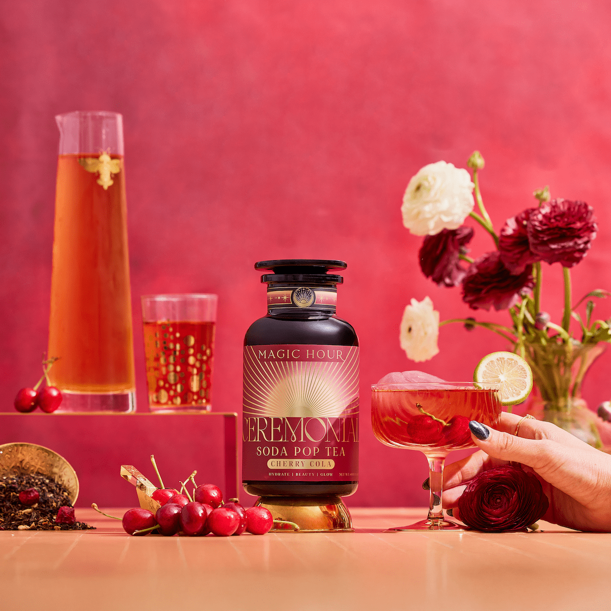 A bottle of Cherry Cola Black Tea: Ceremonial Soda Pop Tea with Sugar &quot;Destroyer&quot; is surrounded by cherries, flowers, a cocktail glass with a lime slice, a tall tea glass, and floating flowers on a pink background.