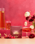 A box of Cherry Cola Black Tea: Ceremonial Soda Pop Tea with Sugar "Destroyer" is surrounded by a teapot, glass, cocktail, cherries, loose tea leaves, and red and white flowers on a pink background.