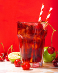 A glass of iced Magic Hour Cherry Cola with two striped straws, garnished with fresh cherries, a lime half, and a whole lime. The drink is placed against a vibrant red background, with some cherries and lime arranged around the glass.